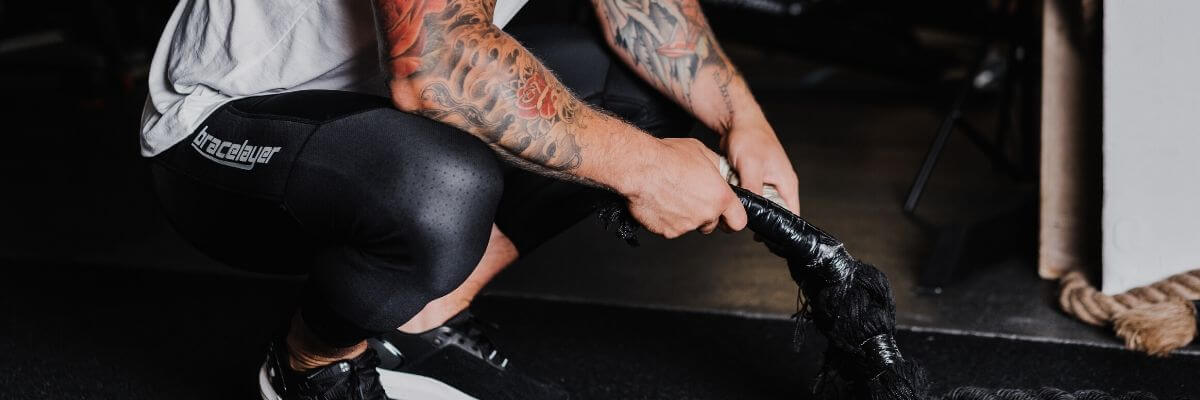An image of a heavily tattooed person kneeling down, wearing Bracelayer's compression leggings with knee support.