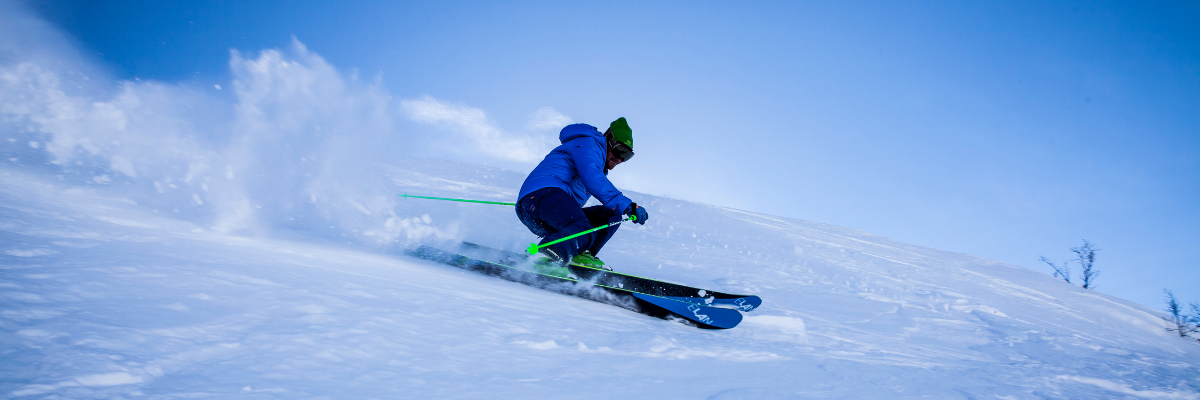 A supportive base layer that is like a hug for your knees! A person skis on a very blue day, wearing a blue ski outfit with bright green poles.