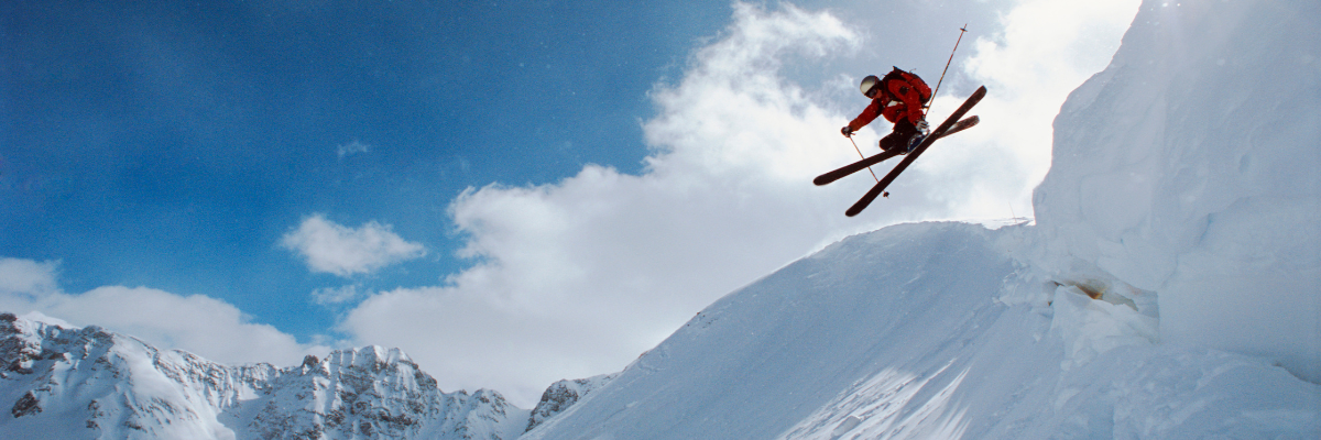 The same trusted knee support brought to you by the KX collection, now a thermal base layer! In this photo, a person is airborne with their skis crossed in a large jump to a lush powdery pile of snow beneath them, with mountains behind them.