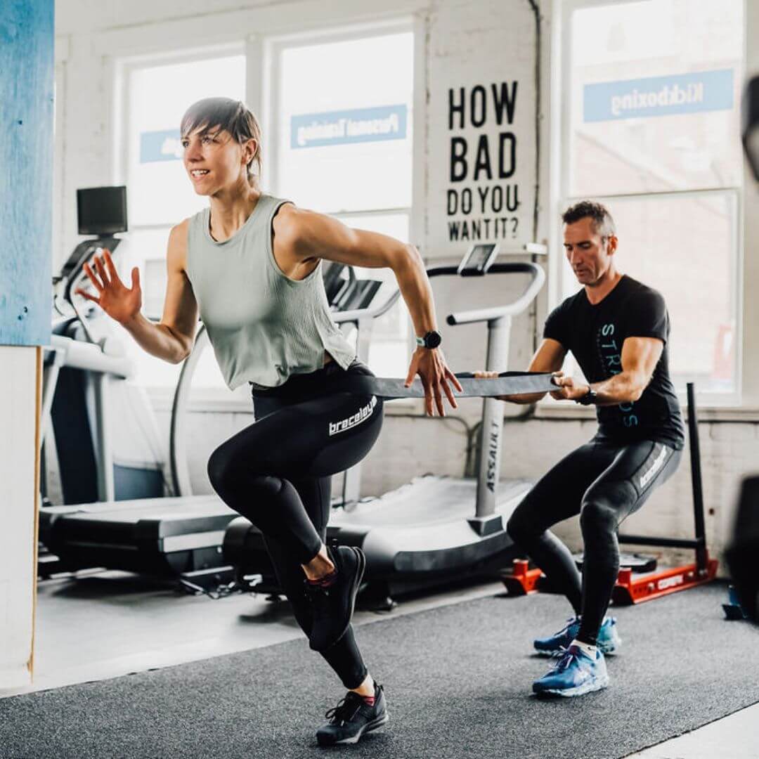 Two people in Bracelayer's Compression Knee Brace Pants are pictured. One has her torso wrapped in a resistance band and is running away from the person behind her for a tough resistance workout!