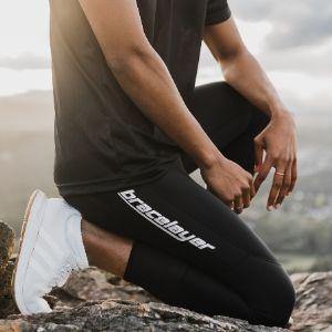 A model in Bracelayer's compression pants with knee support kneels on rocks with gorgeous BC mountains behind him. He is also wearing a black shirt and white sneakers.