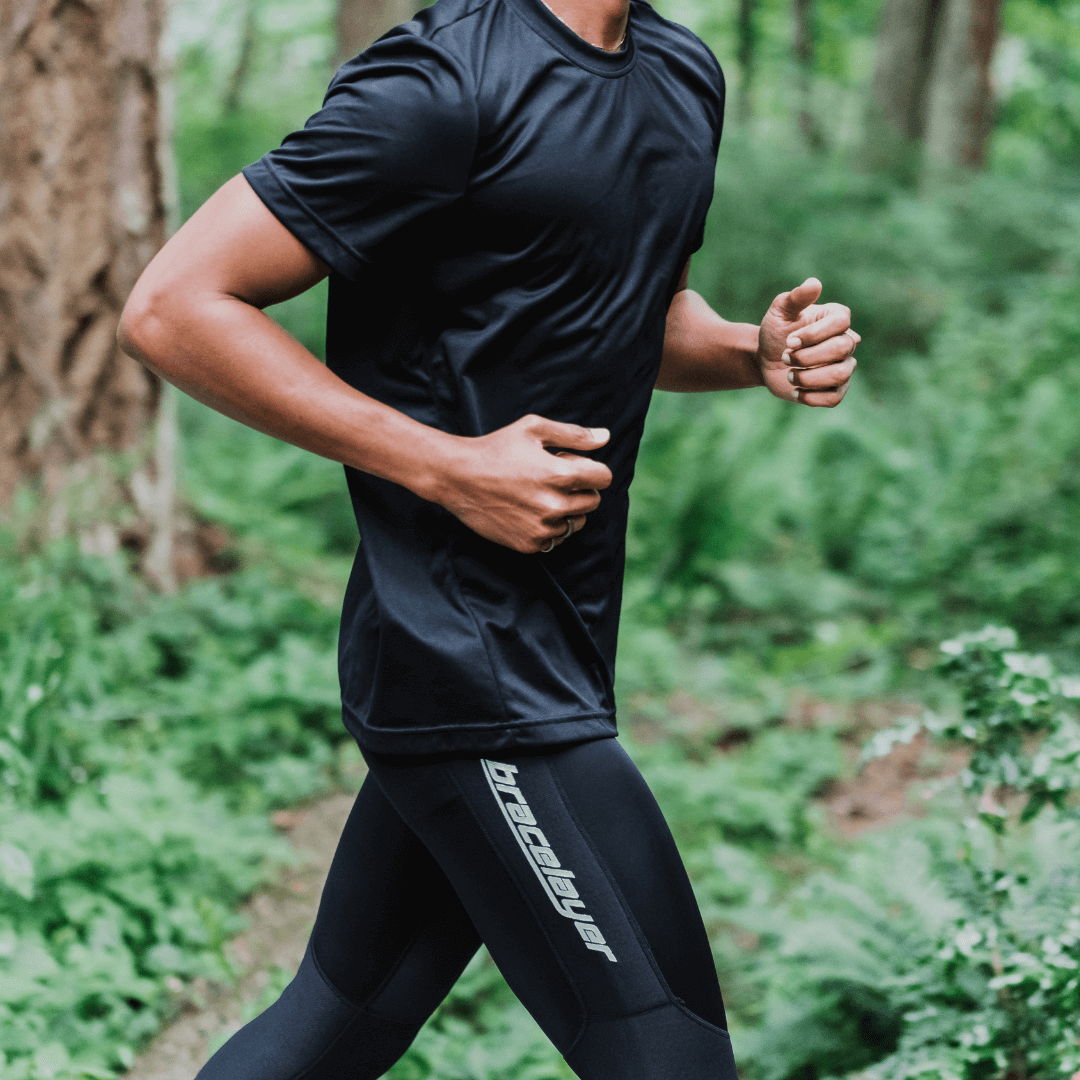 Knee support on the go with Bracelayer's compression exercise pants! In this photo, a young man runs through a lush green forest wearing Bracelayer compression leggings.