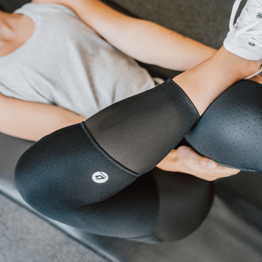 A close up photo of a woman reclined on a yoga mat performing a quadricep stretch, with her right leg crossed over her left, right ankle resting on her knee. She is wearing Bracelayer's KS1 Vent workout pants with knee support for extra stability.