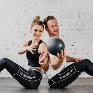 Bracelayer designs innovative compression knee brace tights. Pictured here are two people seated back to back passing a large kettleball between them. They are both wearing Bracelayer's compression pants with knee support for stability and pain relief dur