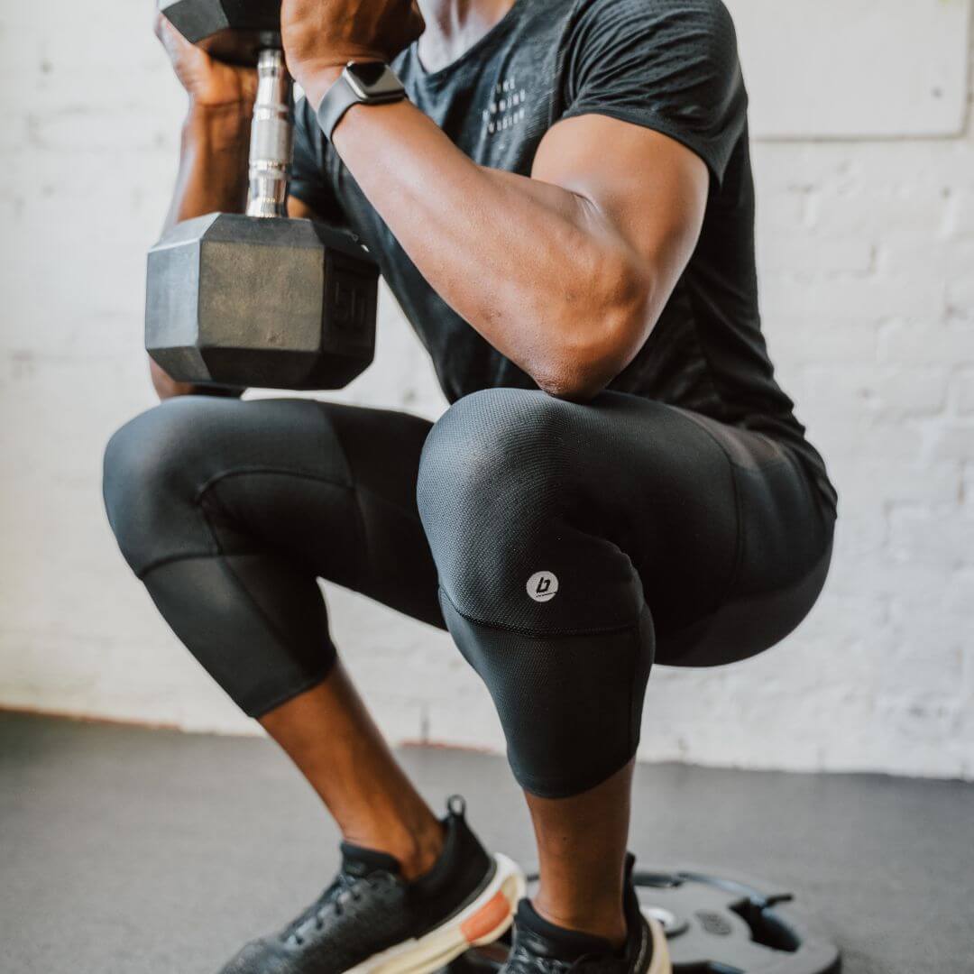 A square image showing a man in Bracelayer's knee brace compression pants, doing a goblet squat while holding a dumbbell. The image is focused closely on his knees.